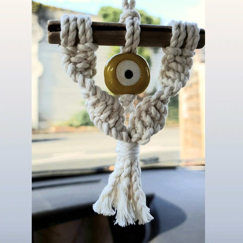 May include: White macrame car charm with a yellow evil eye bead. The charm is hanging from a rearview mirror.