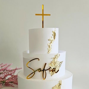 May include: A three-tiered white cake decorated with gold leaf accents and a gold cross on top. The name "Sofia" is written in gold script on the middle tier.