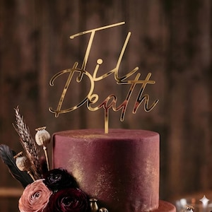 Puede incluir: Un adorno dorado para pastel que dice "Til Death" en una fuente cursiva, colocado encima de un pastel burdeos con detalles dorados. El pastel está decorado con flores rojas oscuras y rosas y otros elementos decorativos.