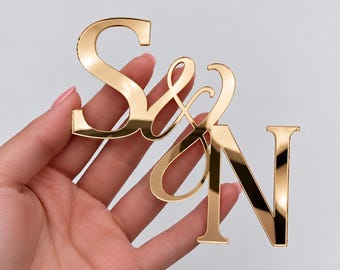 May include: A gold-colored reflective acrylic cutout featuring the letters S and N connected by an ampersand symbol. The letters are rendered in a serif typeface with a smooth, polished surface that creates a mirror-like finish.