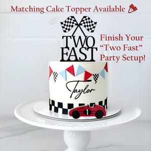 May include: A white cake with a racing theme, featuring a black and white checkered pattern, red and blue pennants, and a red race car. The cake is topped with a black "Two Fast" cake topper and the name "Taylor".