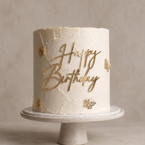 Może przedstawiać: Cylindryczny tort z teksturowanym lukrem w kolorze złamanej bieli, ozdobiony małymi złotymi wycinankami motyli oraz złotym napisem Happy Birthday, umieszczony na jasnej, okrągłej podstawie do serwowania ciast.