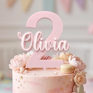 May include: A light pink cake topper featuring the number two with the name Olivia written in white script across the center. The topper is positioned on a frosted cake decorated with light pink flowers, small white pearls, and beige macarons.