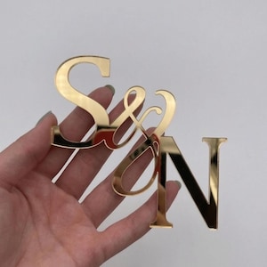 May include: Gold-colored acrylic letters spelling "S&N". The letters are in a cursive font, with the ampersand connecting the "S" and "N". The letters are held in a hand against a white background.