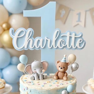 May include: A cake topper with the name "Charlotte" and the number "1". The cake is decorated with an elephant, a teddy bear, and balloons. The cake has blue and white frosting. The cake is decorated with pastel colors.