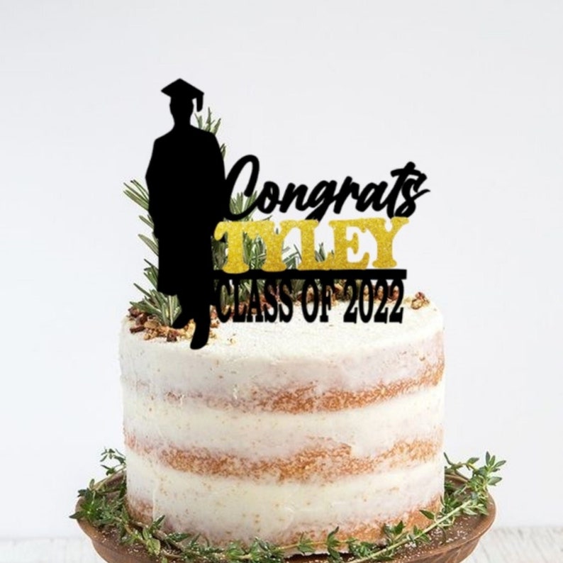 May include: Black and gold graduation cake topper with a silhouette of a graduate, the text "Congrats Tiley Class of 2022", and green foliage.