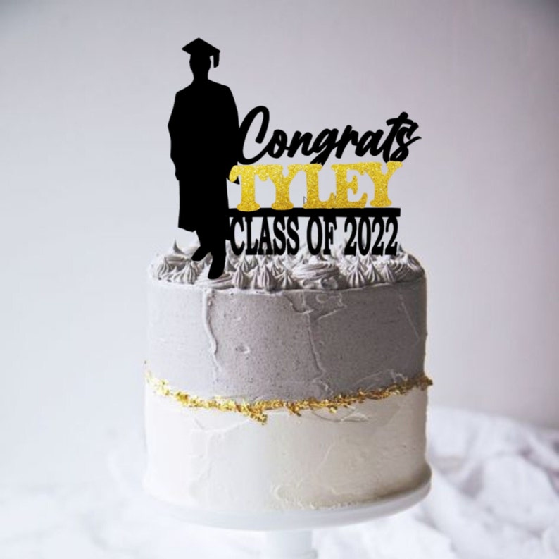 May include: Black silhouette of a graduate with a cap and gown, a gold glitter cake topper that says "Congrats Tyley Class of 2022" on a gray and white frosted cake.