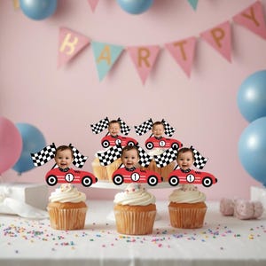 May include: Cupcakes with white frosting and red race car toppers, each with a baby's face and a checkered flag. The background includes a pink wall, balloons, and a banner that says "HAPPY".