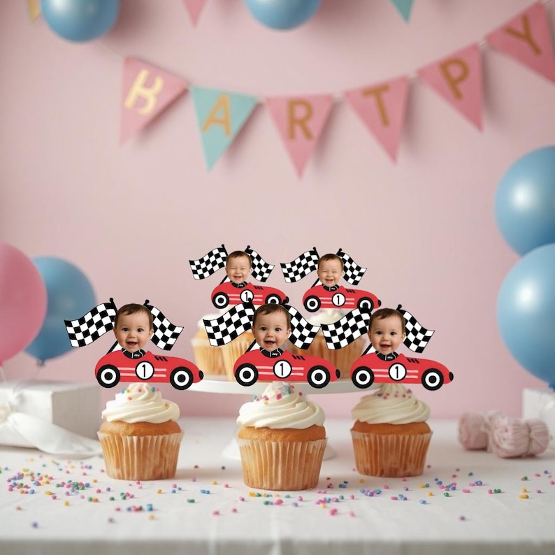 May include: Cupcakes with white frosting and red race car toppers, each with a baby's face and a checkered flag. The background includes a pink wall, balloons, and a banner that says "HAPPY".