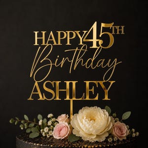 May include: A gold-colored acrylic cake topper featuring the text HAPPY 45TH Birthday ASHLEY in a combination of serif and script fonts. The topper is supported by a thin vertical stake and is positioned above a cluster of light pink and white flowers.