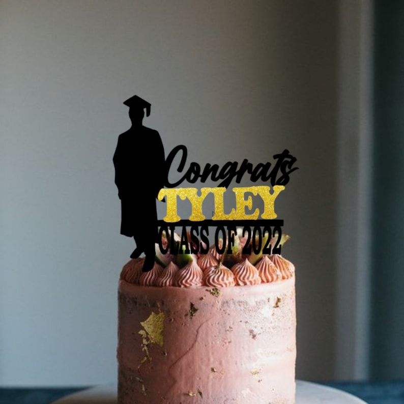 May include: Black silhouette of a graduate with a cap and gown, gold glitter text that reads "Congrats Tyley Class of 2022" on a pink frosted cake.