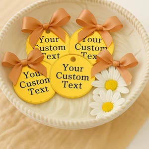 Puede incluir: Primer plano de un cuenco blanco con varias etiquetas de regalo amarillas con lazos marrones. Cada etiqueta tiene el texto "Your Custom Text" en negro. Dos margaritas blancas con centros amarillos también están en el cuenco.