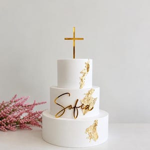 May include: A three-tiered white cake decorated with gold leaf accents. The middle tier features a gold script name topper reading Sofia, and the top tier is topped with a gold cross. The cake has a smooth matte finish on all three levels.