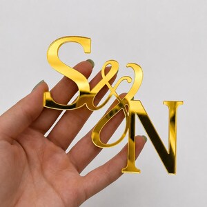 May include: A reflective gold-colored acrylic monogram featuring the letters S and N connected by an ampersand symbol. The letters are rendered in a bold, serif-style font with a smooth, mirror-like finish that catches light across the surface.