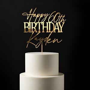 Puede incluir: Un adorno para pastel de color dorado con el texto Happy 60th BIRTHDAY Kayden en una mezcla de letras cursivas y de molde. El adorno está insertado en un pastel blanco de dos niveles y presenta un acabado metálico reflectante.