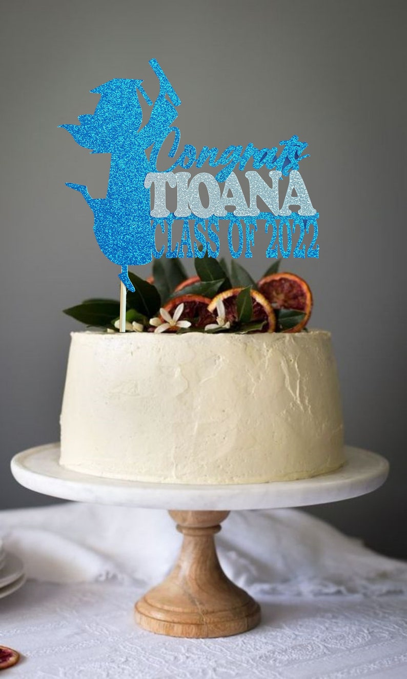 May include: A white cake with a blue and silver glitter graduation cap topper that says "Congrats Tiana Class of 2022". The cake is decorated with orange slices and green leaves.