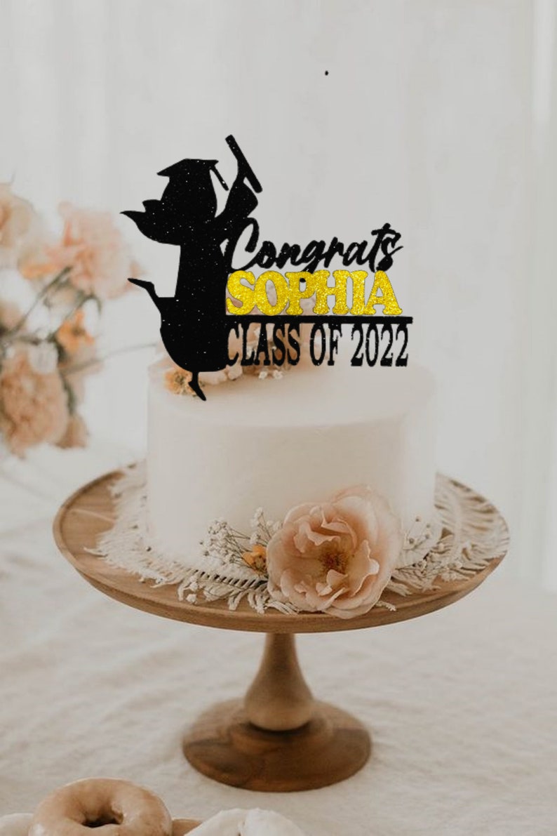 May include: A white cake with a black and gold glitter graduation topper that says "Congrats Sophia Class of 2022". The cake is on a wooden cake stand with a lace trim and a pink rose.