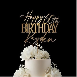 May include: A gold-colored cake topper with the words "Happy 60th Birthday Kayden" in elegant script and block letters. The topper is placed on a white cake decorated with white flowers and greenery against a black background.