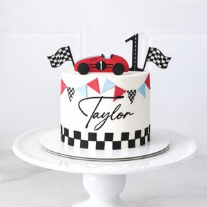 May include: A white cake decorated with a red race car, checkered flags, and the number 1. The cake has a banner of red and blue triangles and the name "Taylor" in script. The base and top of the cake are decorated with a black and white checkered pattern.