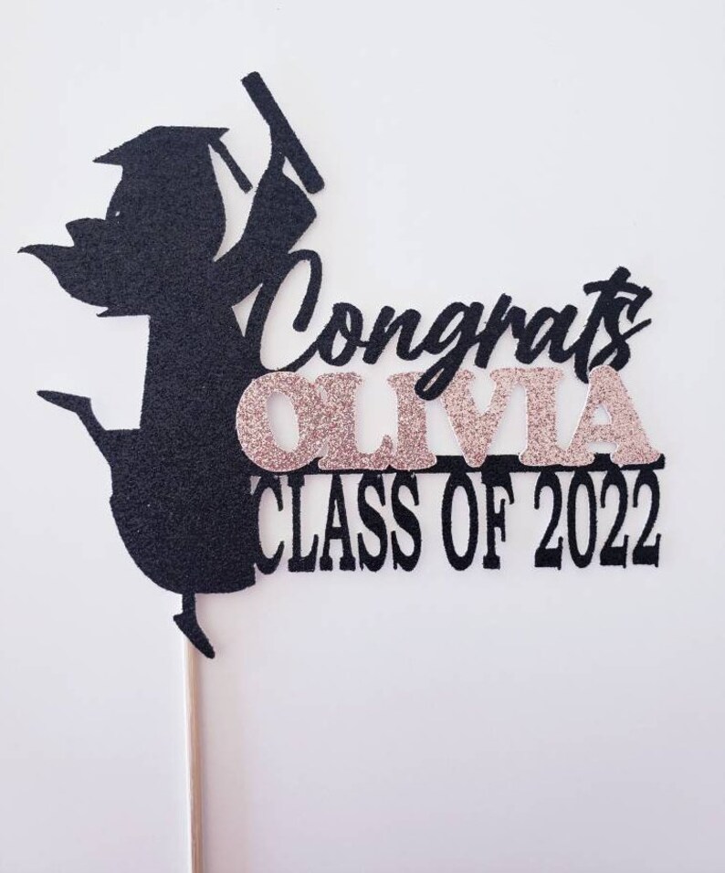 May include: Black graduation cake topper with a silhouette of a person wearing a cap and gown, holding a diploma. The topper says "Congrats Olivia Class of 2022" in black and pink glitter.