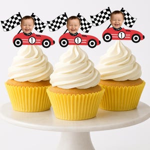 May include: Three cupcakes with white frosting, each topped with a red race car and a baby's face. Checkered flags are positioned behind each car. The cupcakes sit in yellow liners on a white cake stand.