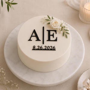 May include: A round white cake topped with a cluster of white roses and green leaves. The cake features black lettering with the initials A and E separated by a vertical line, and the date 8.26.2026 printed in black below the initials.