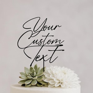 Op de afbeelding: Een zwarte taarttopper met de cursieve tekst Your Custom Text, geplaatst boven een lichtgroene vetplant en een witte dahlia. De topper heeft een dunne verticale steel die naar beneden wijst om in de bovenkant van een taart te steken.