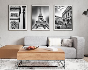 May include: Three black and white framed photographs of Parisian landmarks, including the Eiffel Tower, the Arc de Triomphe, and a street scene. The photographs are hanging on a white wall above a gray sectional sofa and a wooden coffee table.