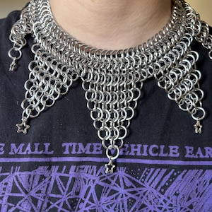 May include: A silver chainmail necklace with a star charm at the bottom of each point. The necklace is worn by a person with a black shirt.