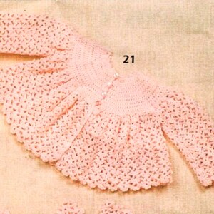 May include: A pink crocheted baby cardigan with long sleeves and a button closure. The cardigan has a scalloped edge and a delicate pattern.