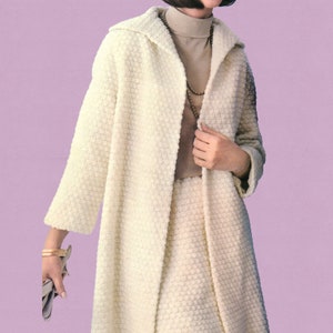 May include: A cream-coloured, textured coat and skirt set. The coat has a wide collar and long sleeves. The skirt is knee-length. The outfit is worn over a tan polo neck and accessorised with a gold necklace and bracelet. A grey clutch is held in the hand.