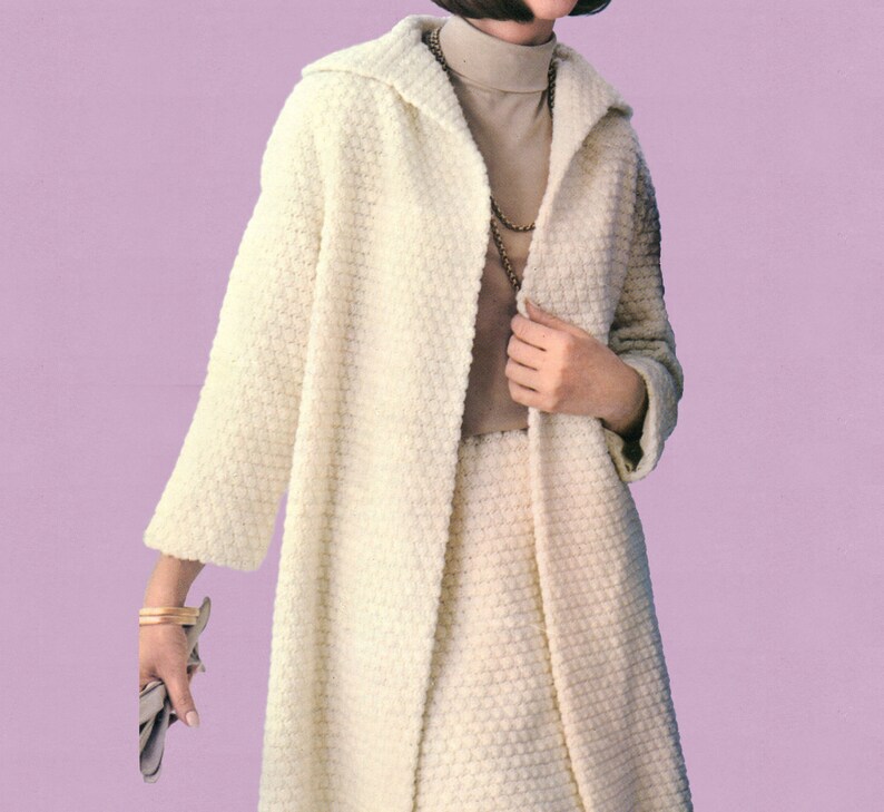 May include: A cream-coloured, textured coat and skirt set. The coat has a wide collar and long sleeves. The skirt is knee-length. The outfit is worn over a tan polo neck and accessorised with a gold necklace and bracelet. A grey clutch is held in the hand.