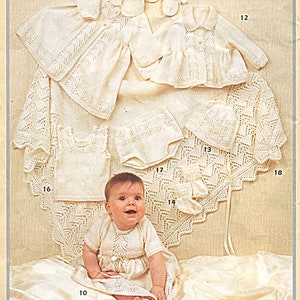May include: A set of white crocheted baby clothes, including a dress, a jacket, a bonnet, booties, and a blanket. The set is laid out on a white satin surface.