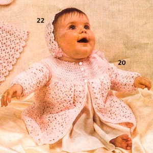 May include: A pink crocheted baby dress with a matching bonnet. The dress has a button closure and a ruffled hem. The baby is wearing a white slip underneath the dress.