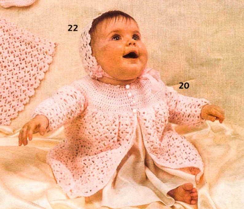 May include: A pink crocheted baby dress with a matching bonnet. The dress has a button closure and a ruffled hem. The baby is wearing a white slip underneath the dress.