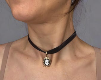 May include: A black velvet choker necklace featuring a central oval pendant. The pendant has a gold-toned ornate frame surrounding a black background with a white profile cameo silhouette of a person facing to the left, suspended by a gold ring.