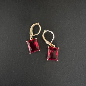 May include: A pair of gold-colored leverback earrings, each featuring a rectangular, deep red gemstone. The earrings are displayed against a dark background, highlighting the contrast between the gold and the red stones.