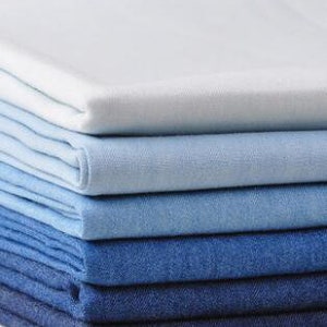 May include: A stack of denim fabric in various shades of blue, from light to dark. The fabric is folded neatly and arranged in a gradient pattern.