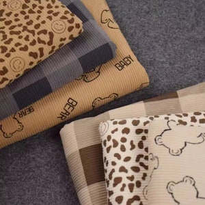 Könnte beinhalten: Ein Stapel aus fünf verschiedenen Stoffmustern mit verschiedenen Mustern. Das oberste Muster ist ein brauner und beigefarbener Leopardenprint. Das zweite Muster ist ein grau-weißes Karomuster. Das dritte Muster ist ein beiger Stoff mit einem wiederholten Druck des Wortes "BABY". Das vierte Muster ist ein beiger Stoff mit einem wiederholten Druck des Wortes "BEAR". Das unterste Muster ist ein beige-braunes Karomuster.