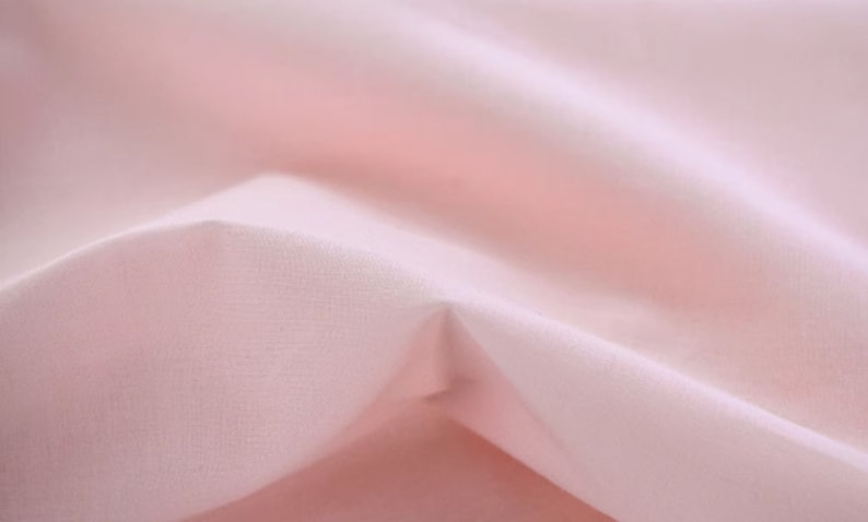 May include: A close-up of a soft, pale pink fabric with a smooth, even texture. The fabric is folded and draped, creating a gentle, flowing effect.