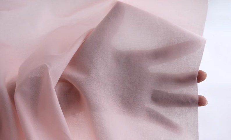 May include: A pale pink sheer fabric, draped and held in a hand. The fabric is soft and delicate, with a subtle sheen.