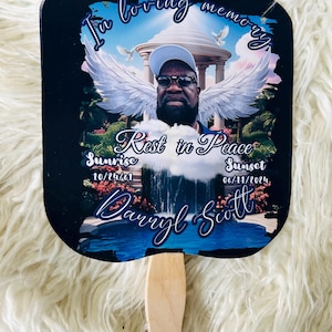 May include: A memorial fan with a photograph of a man wearing sunglasses and a cap, with angel wings superimposed. The fan includes the text "In loving memory", "Rest in Peace", and the dates "10/24/61" and "06/11/2024". The fan has a wooden handle.