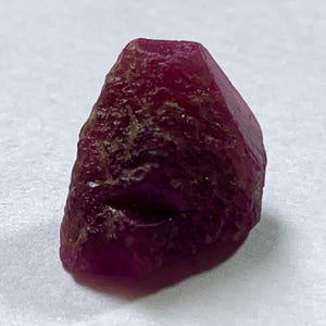 May include: A rough-cut, deep red ruby gemstone with a faceted top and a textured, uneven surface. The ruby is a rich, dark red color and appears to be a natural, unpolished stone. The gemstone is set against a white background.