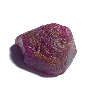 Peut inclure: Une pierre précieuse brute de rubis. Le rubis est d'un rouge profond et riche, avec des reflets dorés et bruns. La pierre a une forme naturelle et irrégulière, sur fond blanc.