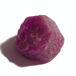 May include: A rough, uncut ruby gemstone, displaying a deep magenta color. The natural stone has a textured surface and irregular shape, casting a shadow on the white background. This gemstone is suitable for jewelry making or collecting.