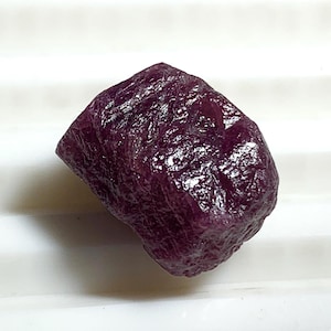 May include: A rough, uncut ruby gemstone with a deep, rich burgundy color. The surface of the stone has a textured, uneven appearance. The ruby is displayed against a white background.