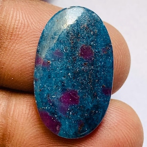 May include: An oval-shaped gemstone with a teal base color and scattered deep red inclusions. The polished surface reflects light, highlighting the contrasting colors and textures of the natural stone. The gemstone is held in a hand.