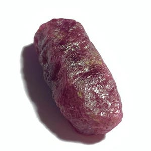 Könnte beinhalten: Ein roher, ungeschliffener Rubin-Edelstein. Der Rubin hat eine tiefe, satte rote Farbe mit einer leicht rauen, strukturierten Oberfläche. Der Edelstein ist länglich und zylindrisch geformt und hat ein natürliches, unpoliertes Aussehen.