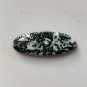 Op de afbeelding: Ovale edelsteen met een witte basis en ingewikkelde patronen van donkergroen, die doen denken aan mos of bladeren. Het gepolijste oppervlak reflecteert het licht en benadrukt de natuurlijke kleur- en textuurvariaties. De edelsteen staat tegen een neutrale achtergrond.