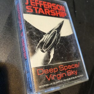 May include: A cassette tape with a black and white image of a spaceship on the front. The text "Jefferson Starship" is in red letters at the top of the tape. The text "Deep Space/Virgin Sky" is in red letters at the bottom of the tape. The text "Recorded Live on Earth at the House of Blues, Hollywood" is in smaller red letters below the title.